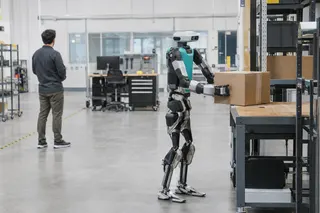Humanoid robot lifts a cardboard box from a warehouse shelf as a person stands in the background.