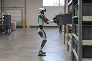 Humanoid robot lifts a plastic bin onto a warehouse shelf, surrounded by storage racks and industrial shelving.
