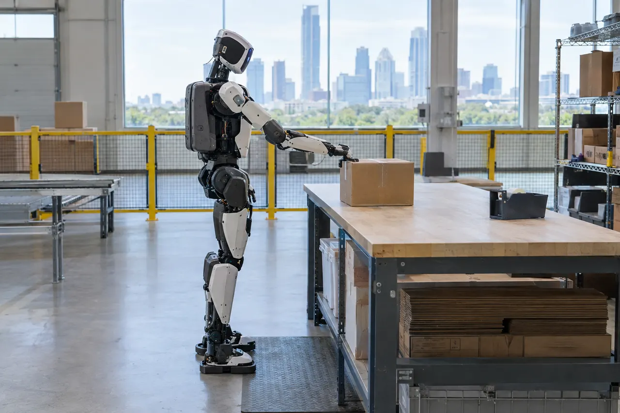 Humanoid robot stands beside a warehouse table, reaching for a cardboard box with a city skyline visible through large windows.