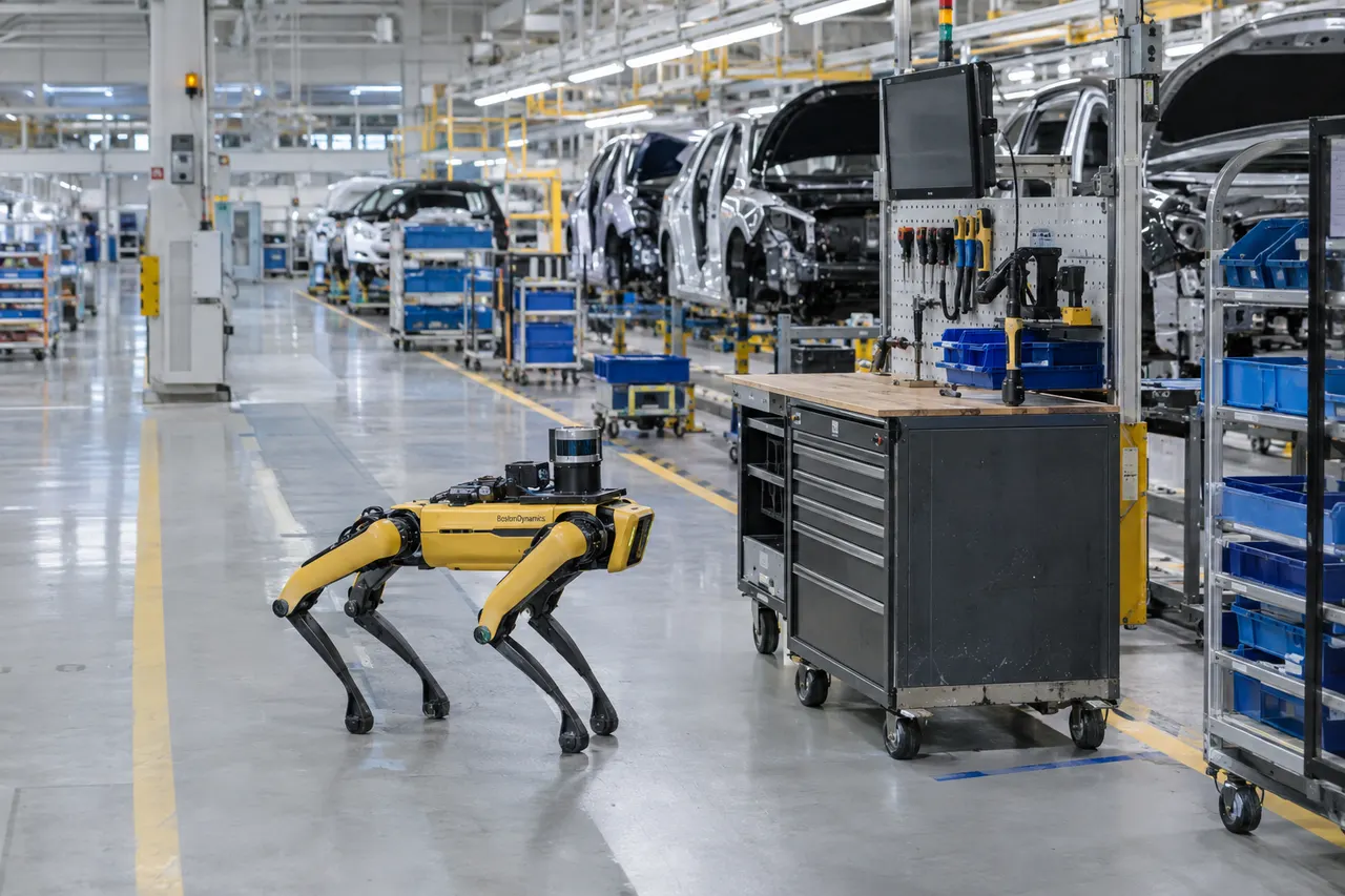 Yellow Boston Dynamics robot dog patrols an automotive factory floor lined with car bodies and industrial equipment.