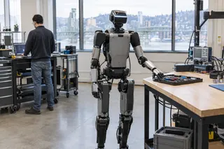 Humanoid robot stands beside a workbench in a lab, with a person working at a nearby workstation in the background.