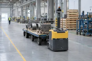 Autonomous tugger hauls metal parts on a cart through a spacious, well-lit industrial warehouse with high ceilings.
