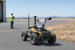 Wheeled firefighting robot with mounted equipment on airport tarmac, operator in safety vest standing in the background.