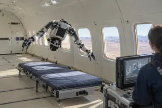 Humanoid robot hovers above padded platform inside aircraft while a person monitors controls on a screen nearby.