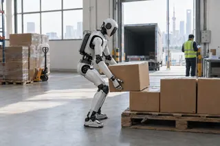 Humanoid robot lifts a cardboard box onto a pallet inside a warehouse as a worker stands near an open loading dock.