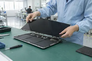 Technician in a lab coat installing a thin display panel onto a disassembled MacBook on a workbench.