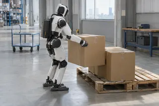 Humanoid robot in a warehouse lifting a cardboard box onto a wooden pallet with more boxes nearby.