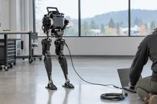 Bipedal research robot connected to a laptop by cable, standing in a bright, modern lab with large windows.