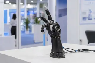 Robotic hand with articulated fingers posed upright on a table in a modern exhibition setting.