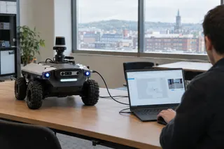 Person uses a laptop to program a wheeled robot in a modern office with large windows overlooking a cityscape.