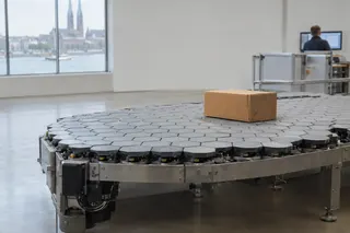 Cardboard box sits on a hexagonal-tiled robotic conveyor in a modern warehouse near large windows with city view.