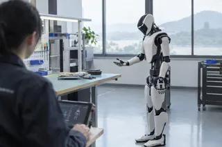 Humanoid robot stands in a bright lab facing a seated person with a tablet, workbench and tools visible in the background.