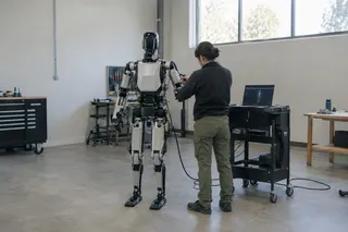 Technician adjusts a humanoid robot in a spacious, well-lit workshop with tools and a laptop on a nearby cart.